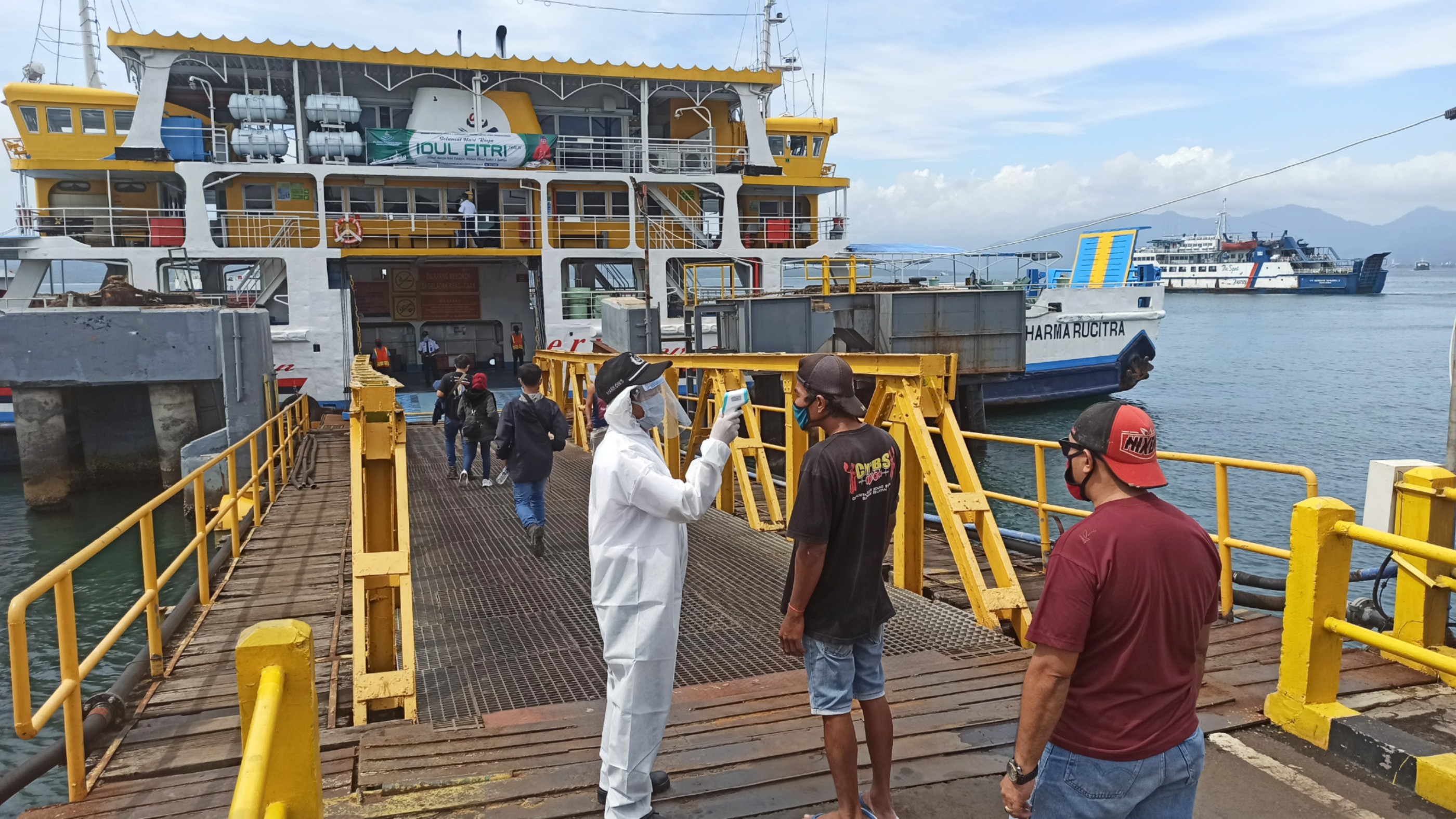 Kru salah satu kapal penyeberangan mengenakan baju Hazmat lengkap dengan face shield, saat memeriksa suhu tubuh calon penumpang yang akan naik ke atas kapal penyeberangan. (Foto: Muh Hujaini/Ngopibareng.id)