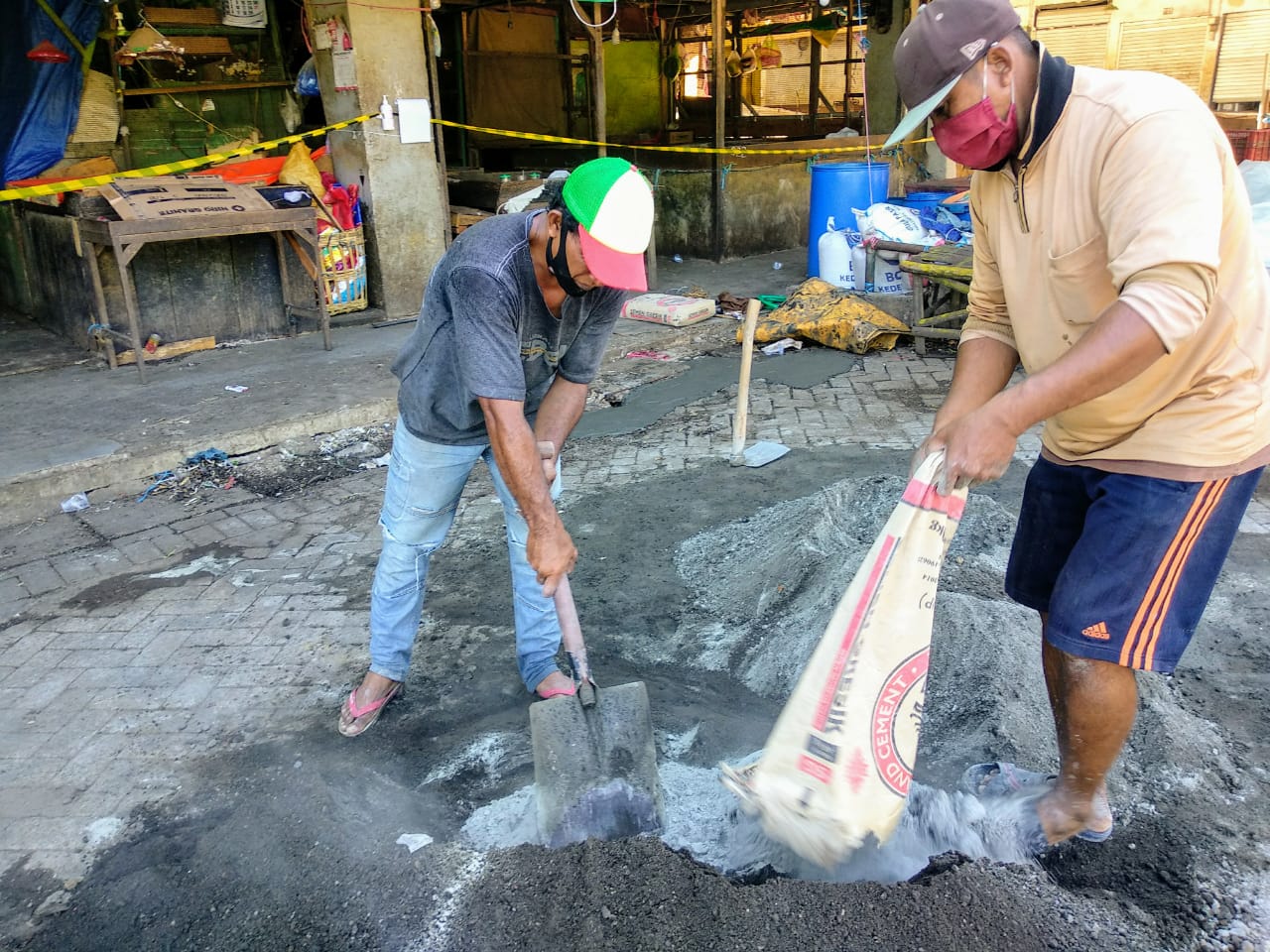 Dua kuli bangunan tampak mencampur semen dengan pasir untuk menambal halaman depan pasar yang rusak. Mereka memakai topi dan masker wajah. (Foto: M.Rizqi/Ngopibareng.id)