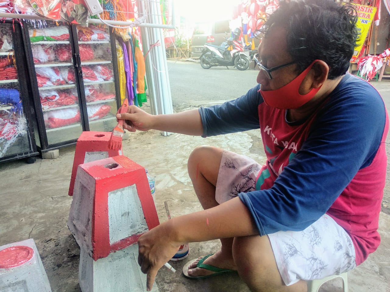 Salah satu penjual akseoris mengecat tiang bendera berwarna merah menggunakan kuas. Pria berkacamata itu memakai masker dengan warna yang sama, dan di tangannya yang lain ia memegang rokok yang dihisapnya (Foto: M.Rizqi/Ngopibareng.id)