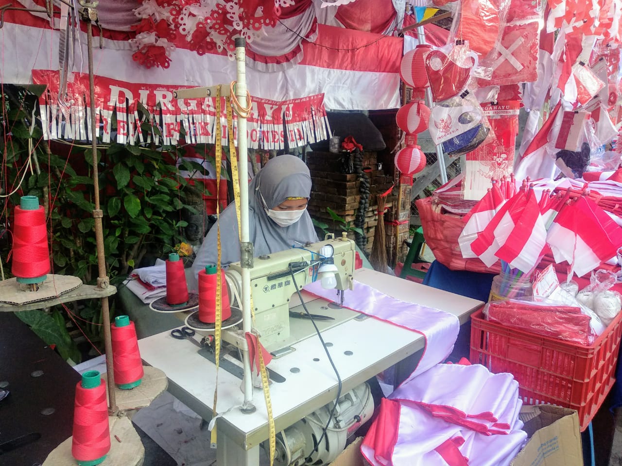 Penjual aksesoris kemerdekaan sedang menjahit bendera menggunakan mesin jahit. Perempuan berkerudung itu tetap mematuhi protokol kesehatan dengan memakai masker berwarna putih (Foto: M.Rizqi/Ngopibareng.id)