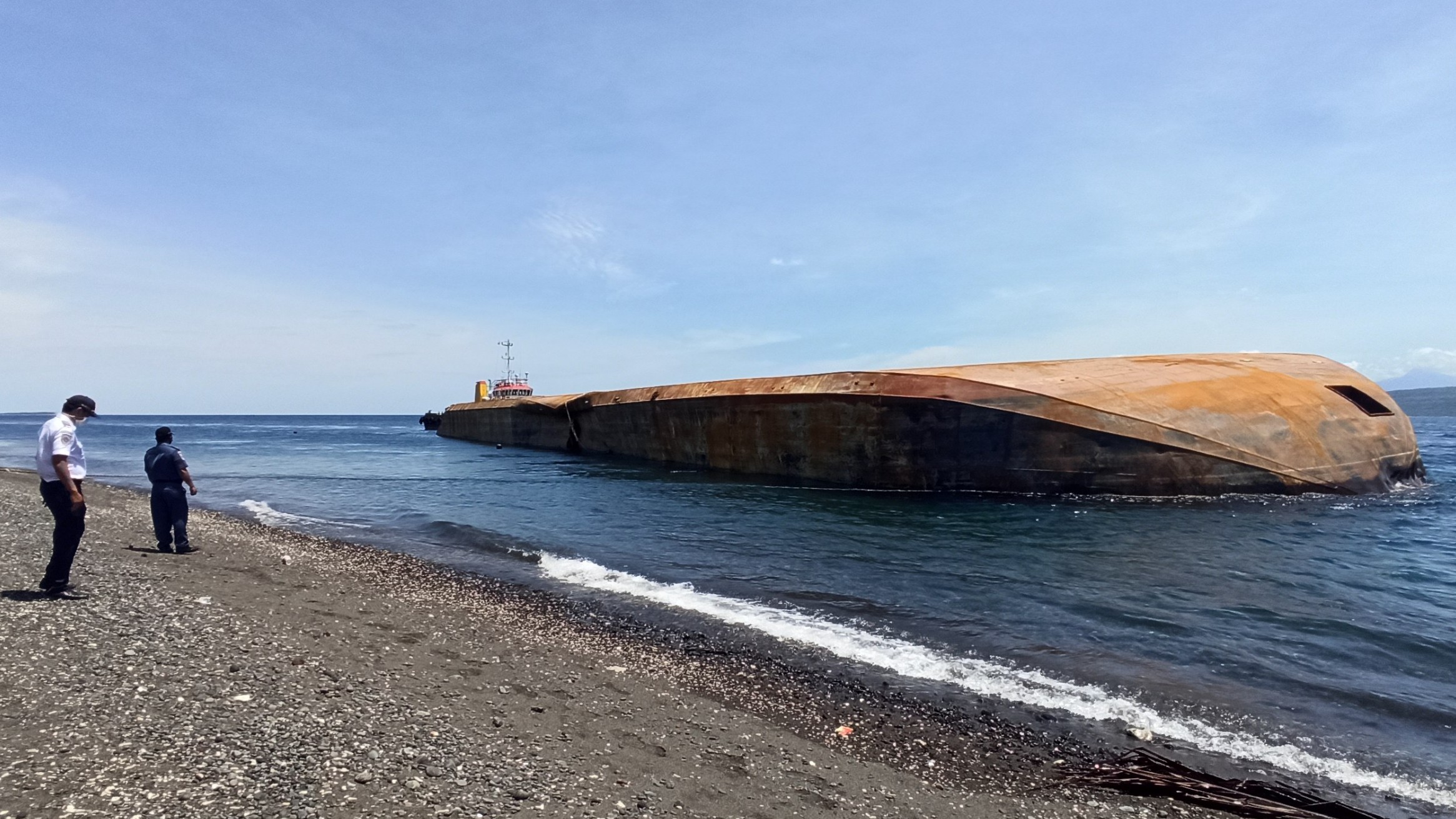 Petugas KSOP Tanjungwangi melihat Kapal tongkang Tan 11 yang masih dalam posisi terbalik di Pantai masuk Desa Bangsring, Kecamatan Wongsorejo (foto:Muh Hujaini/Ngopibareng.id)