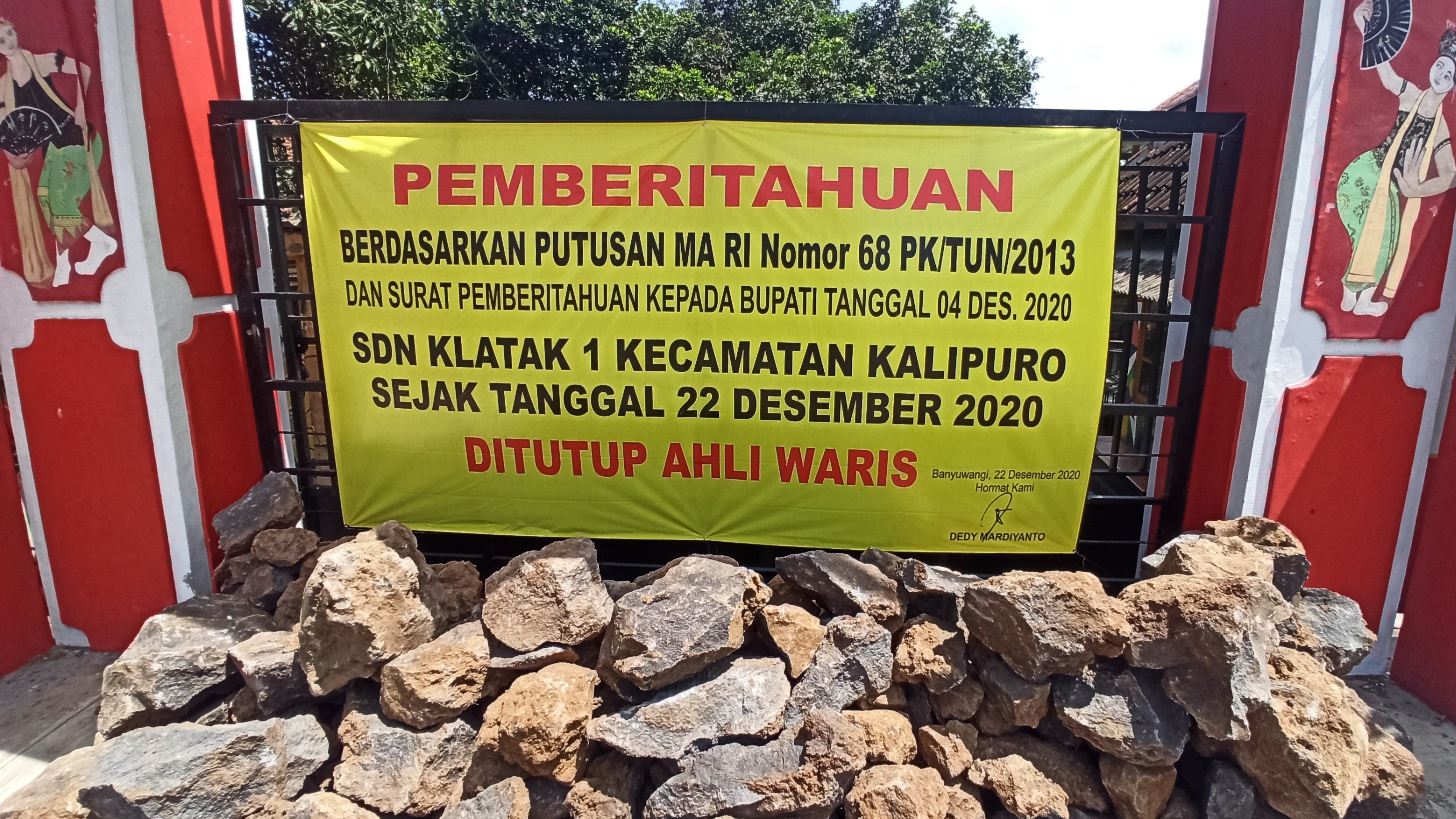 Banner penutupan SDN 1 Klatak dan batu yang ditimbun di depan pintu masuk sekolah ini (foto: Muh Hujaini/Ngopibareng.id)