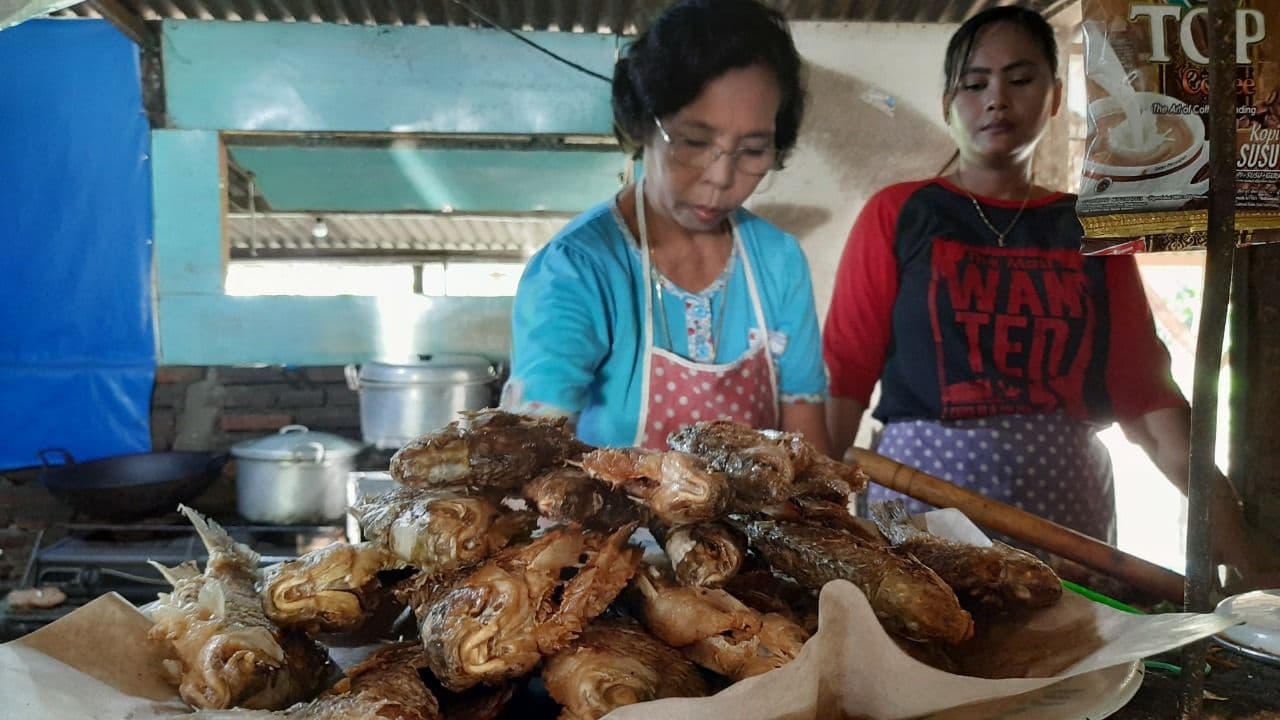 Suciwati, usia 60 tahun, warga Desa Gumulan, Kecamatan Kesamben, saat beraktivitas di warungnya. Olahan ikan yang disajikan Suciwati, diambil langsung dari sungai Brantas di sisi utara warung. (Foto: Mardiansyah Triraharjo/Ngopibareng.id)