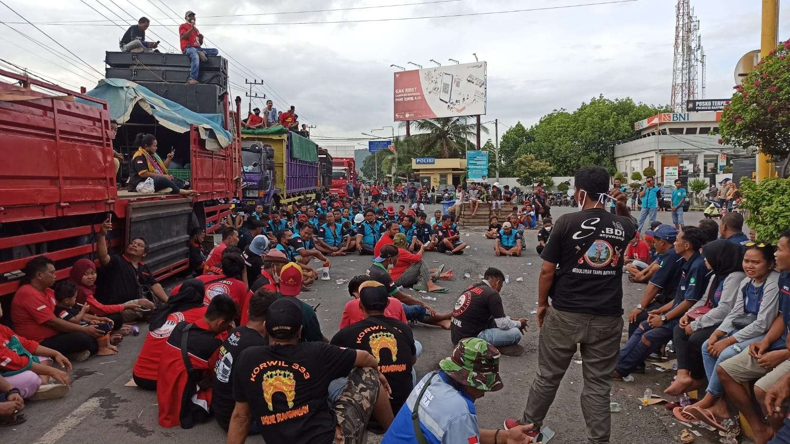 Para sopir logistik mendengarkan orasi dari teman-temannya sambil duduk di jalan depan pintu masuk Pelabuhan Ketapang (foto: Muh Hujaini/Ngopibareng.id)