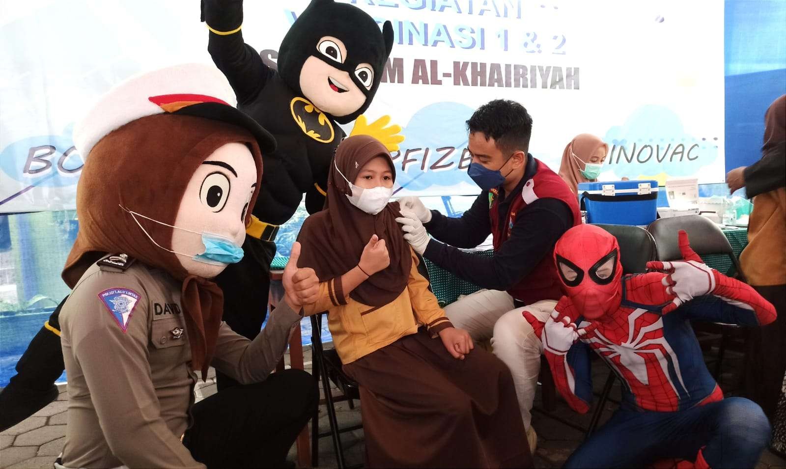 Dua superhero dan maskot Polisi Lalu Lintas menemani seorang siswi yang sedang melakukan vaksinasi (foto:Muh Hujaini/Ngopibareng.id)