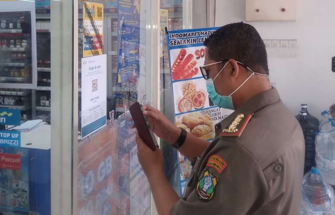 Kepala Satpol PP Bondowoso Slamet Yantoko mencoba barcode Aplikasi PeduliLindungi terpasang depan Toko Indomaret. (Foto: Guido Saphan/Ngopibareng.id)