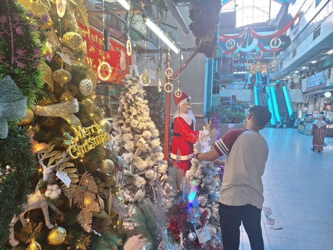 Seorang pegawai gerai pernak-pernik Natal sedang membenahi pohon Natal yang dipajang pada salah satu gerai di Pasar Atom, Surabaya. (Foto: Julianus Palermo/Ngopibareng.id)