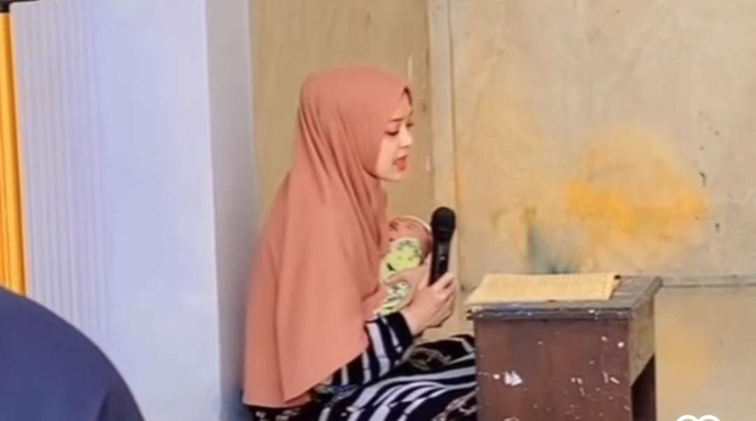 Seorang ibu bersama anaknya di gendongan, mendaras Al-Quran.