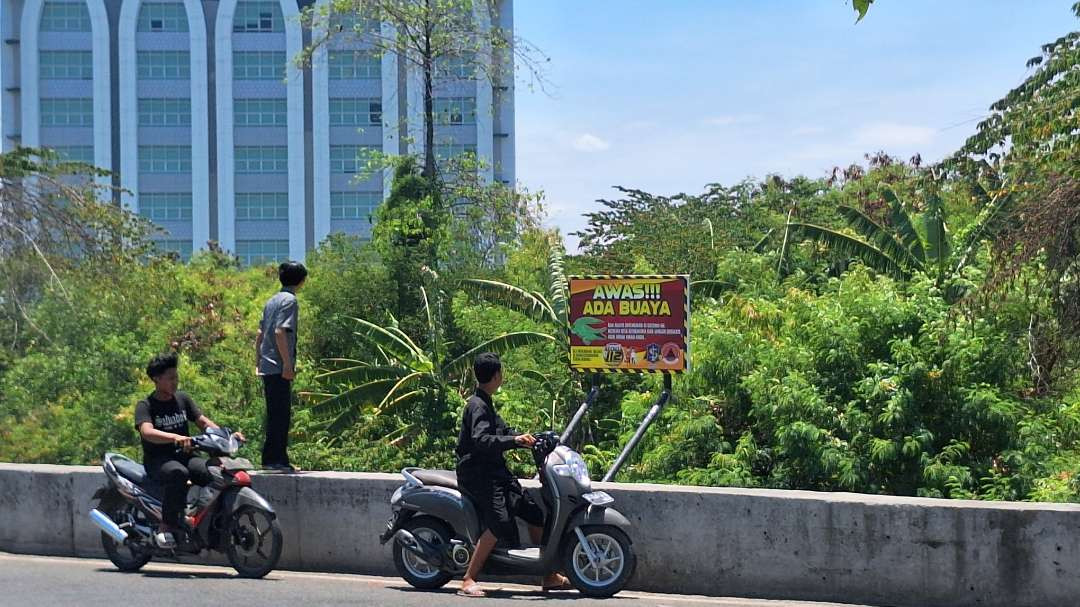 Warga yang melihat dan melintas di dekat papan imbauan peringatan terhadap buaya di Sungai Gunung Anyar dekat Kampus UINSA. (Foto: Julianus Palermo/Ngopibareng.id)